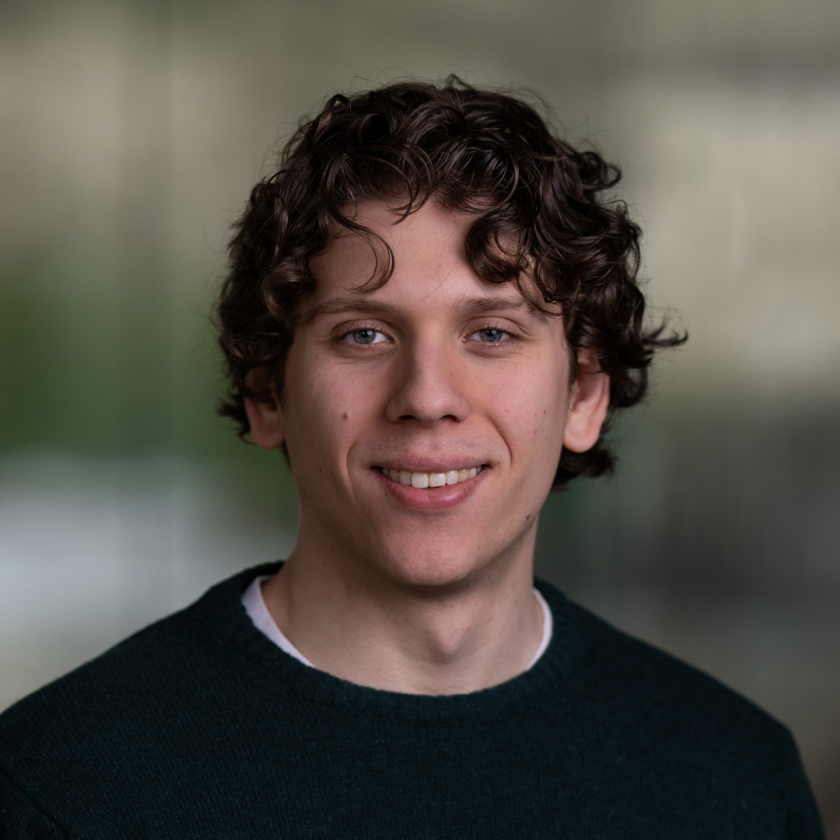 A young man with curly brown hair and a dark green sweater smiles at the camera against a blurred background.