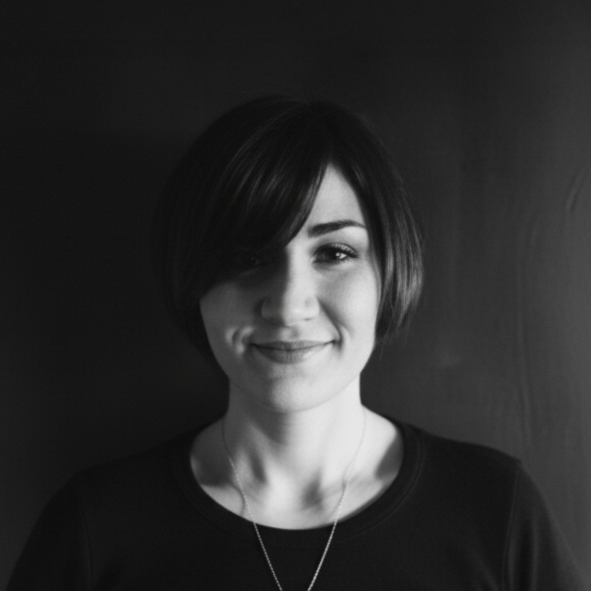 A person with short, dark hair and a side fringe, wearing a dark top and a necklace, stands against a plain dark background.