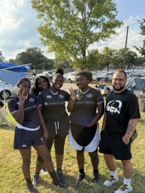 Four young women in matching sports uniforms and a man in a black t-shirt stand together outdoors on grass, posing and smiling for the camera.
