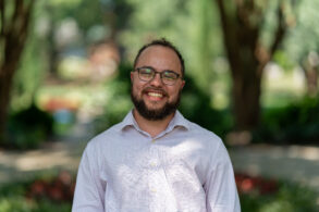 A man with glasses and a beard smiles outdoors, wearing a light-colored button-up shirt. The background is blurred greenery.