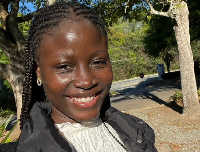 A woman with braided hair smiles outdoors, wearing a black jacket and white top, with trees and sunlight in the background.