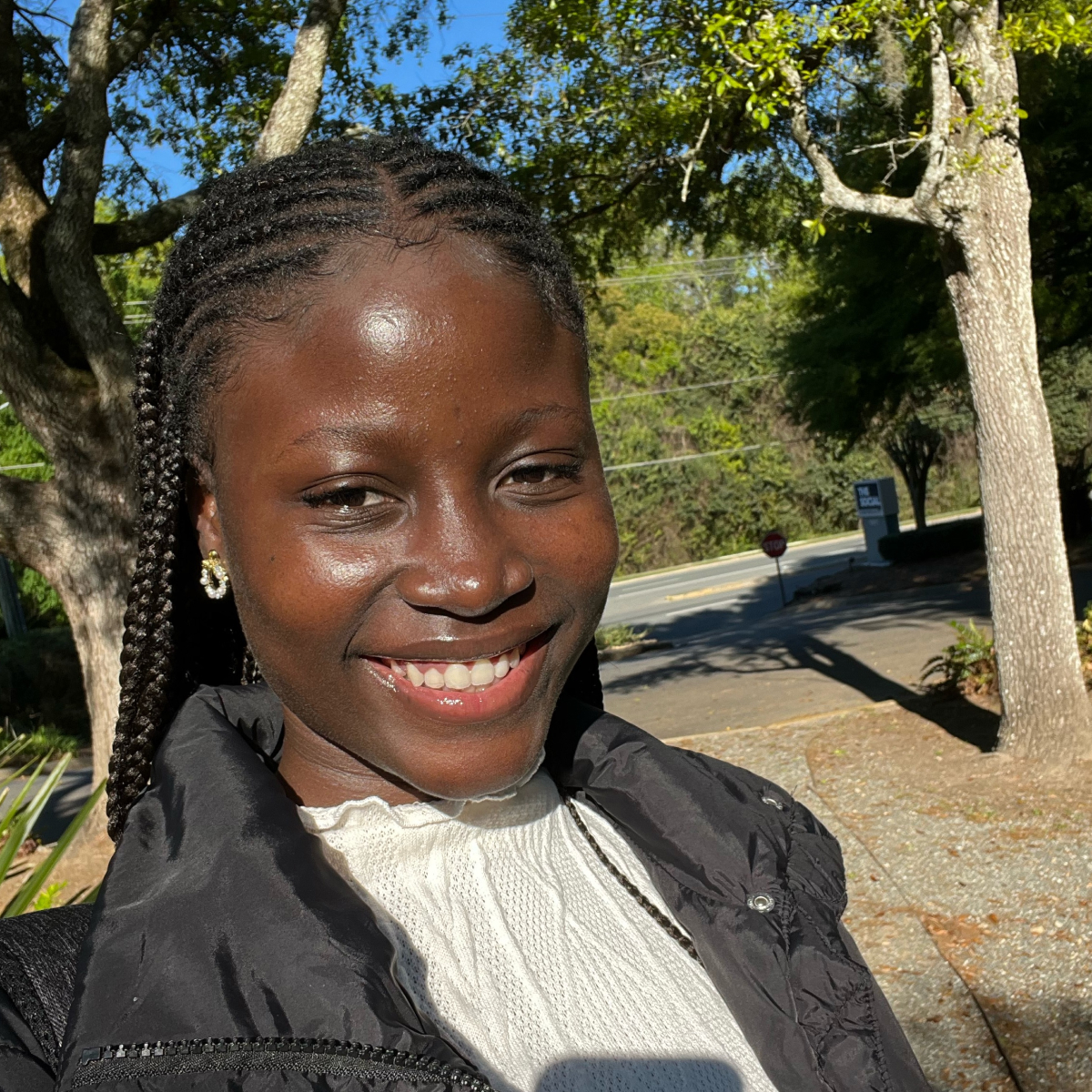A woman with braided hair smiles outdoors, wearing a black jacket and white top, with trees and sunlight in the background.