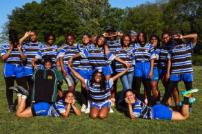 A group of teenage girls in blue and black striped rugby uniforms posing and smiling together on a grassy field with trees in the background.