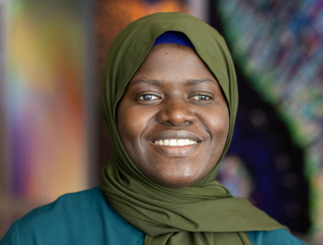 A woman wearing a green hijab and teal top smiles at the camera; colorful, blurred artwork is visible in the background.