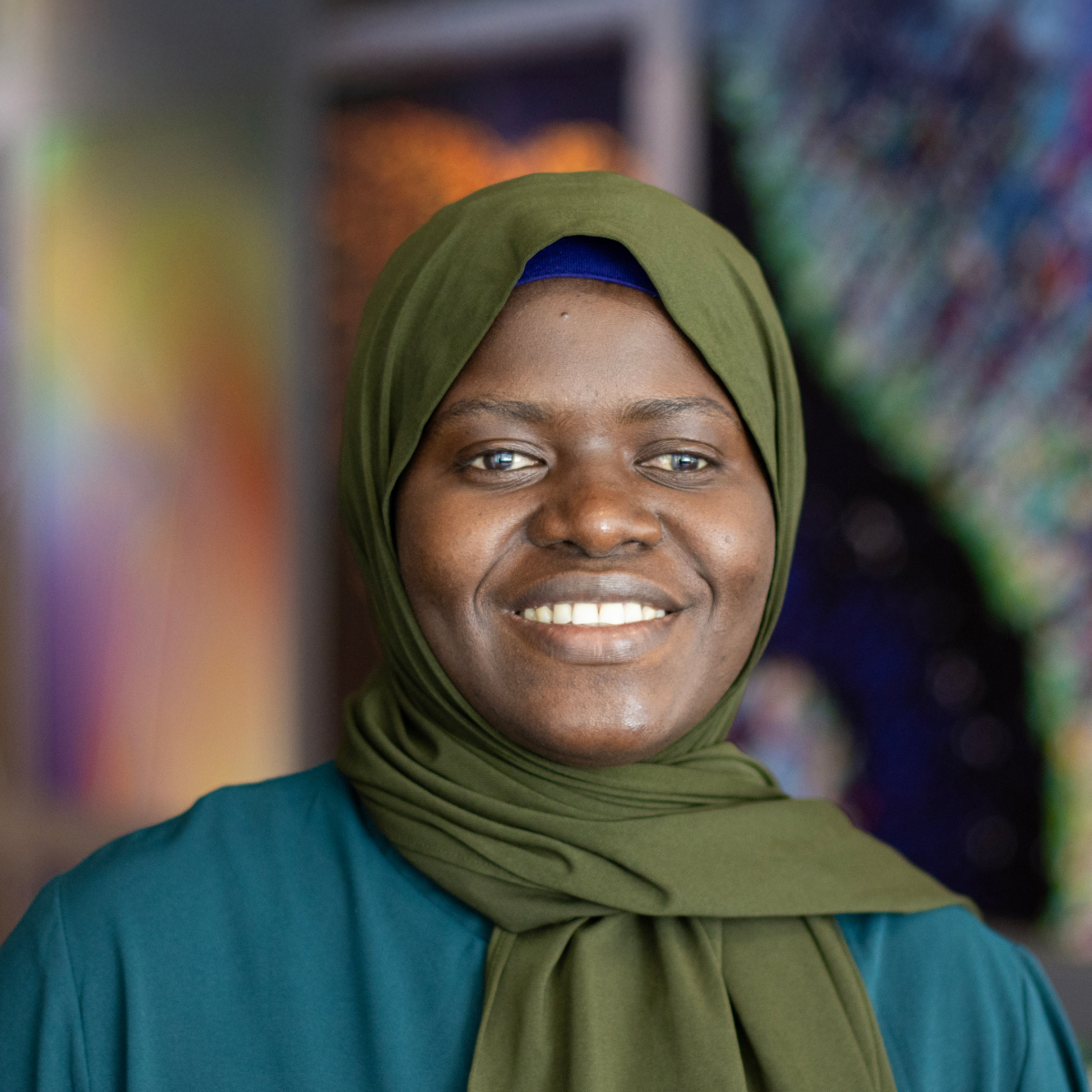A woman wearing a green hijab and teal top smiles at the camera; colorful, blurred artwork is visible in the background.