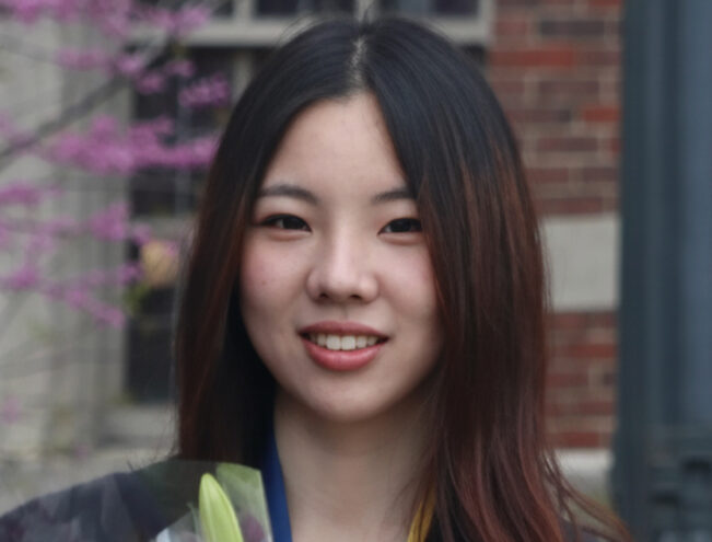 A young woman in graduation attire holds a bouquet of lilies and smiles in front of a brick building with blooming trees.