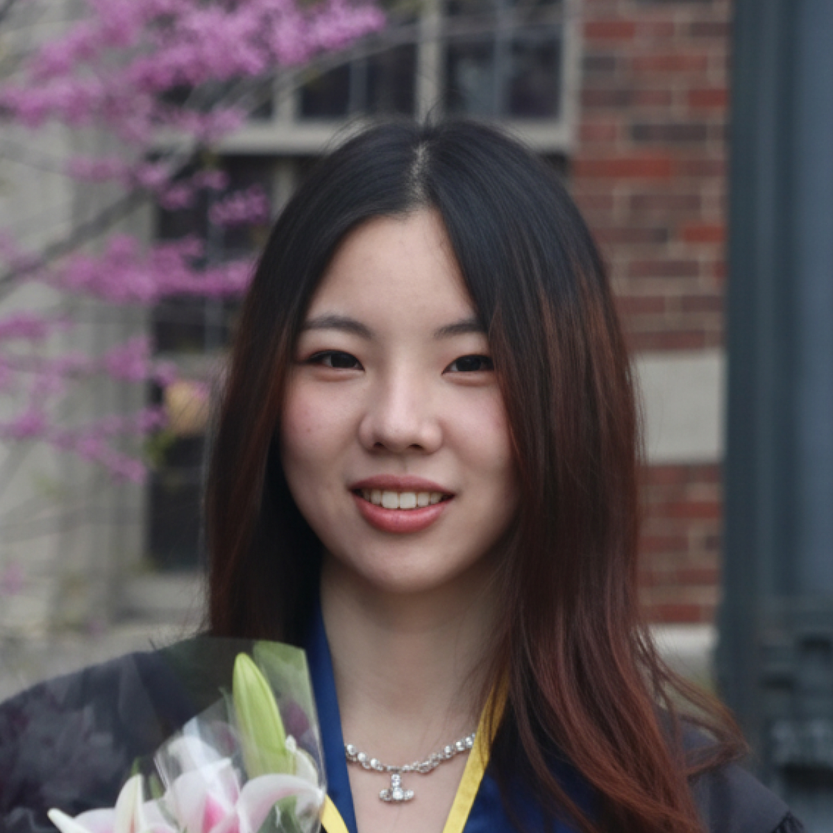 A young woman in graduation attire holds a bouquet of lilies and smiles in front of a brick building with blooming trees.