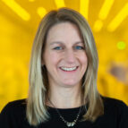 A woman with straight blonde hair and a black top smiles at the camera. The background is yellow and softly blurred with bright light patterns.