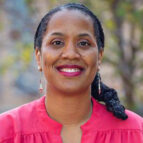 A woman with dark braided hair, wearing a bright pink blouse and drop earrings, smiles at the camera against a blurred outdoor background.