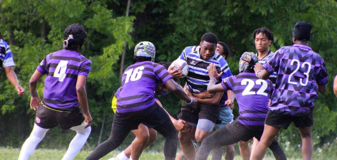 A rugby player in a blue and black striped jersey runs with the ball while four opponents in purple jerseys attempt to tackle him on a grassy field.