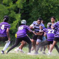 A rugby player in a blue and black striped jersey runs with the ball while four opponents in purple jerseys attempt to tackle him on a grassy field.
