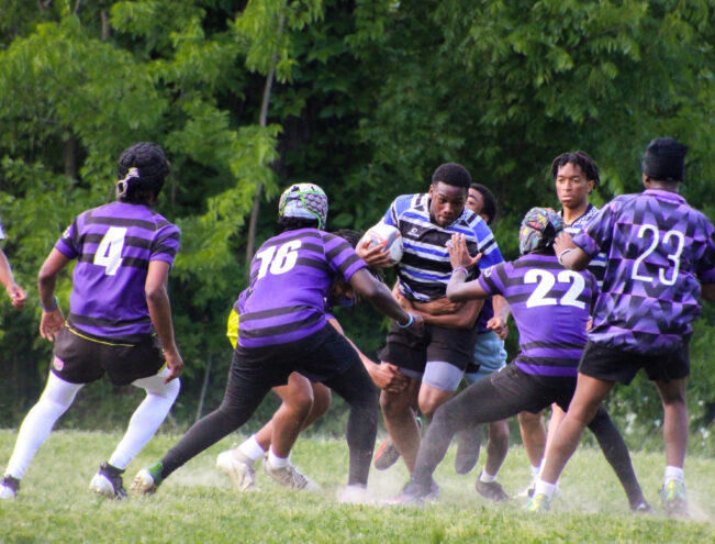 A rugby player in a blue and black striped jersey runs with the ball while four opponents in purple jerseys attempt to tackle him on a grassy field.