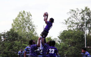 A rugby player is lifted by teammates to catch the ball during a lineout, with opponents and trees in the background.