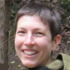 A person with short brown hair smiles outdoors, with greenery and trees in the background.