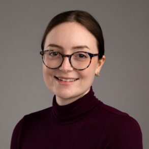 A woman with glasses and brown hair pulled back, wearing a maroon turtleneck sweater, smiles at the camera against a plain gray background.