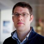 A man with short brown hair and glasses, wearing a navy jacket and light blue shirt, stands indoors with a neutral expression.