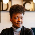 A woman with short curly hair wearing a light turtleneck and dark cardigan stands indoors, smiling slightly with arms crossed.
