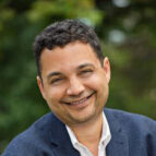 Smiling man with short curly hair wearing a blue blazer and white shirt, outdoors with greenery in the background.
