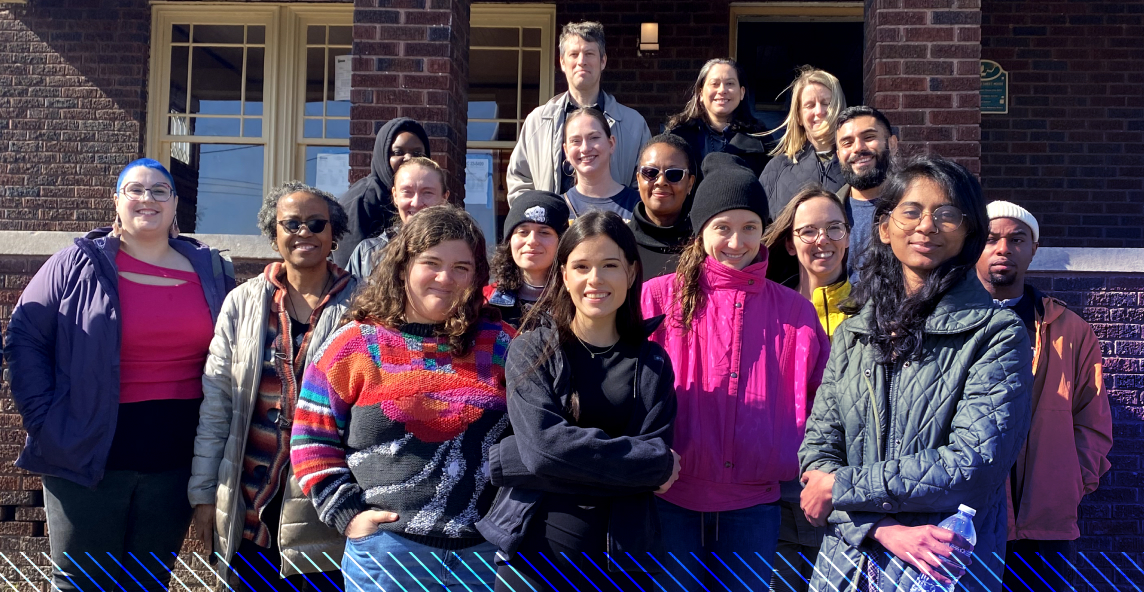 A group of sixteen people stand together outside in front of a brick building, posing for a group photo on a sunny day.