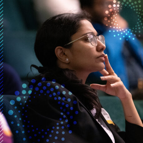 A woman wearing glasses and a suit sits in an audience, resting her chin on her hand and looking thoughtful. Blue decorative dots overlay the image.