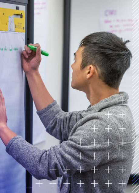 A person in a gray sweater writes on a large sheet of paper attached to a wall with a green marker.