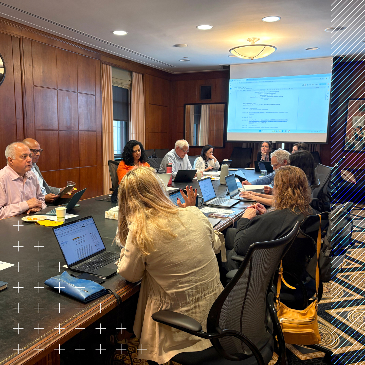 A group of people sit around a conference table with laptops open, engaged in a meeting; a document is projected on a screen at the front of the room.