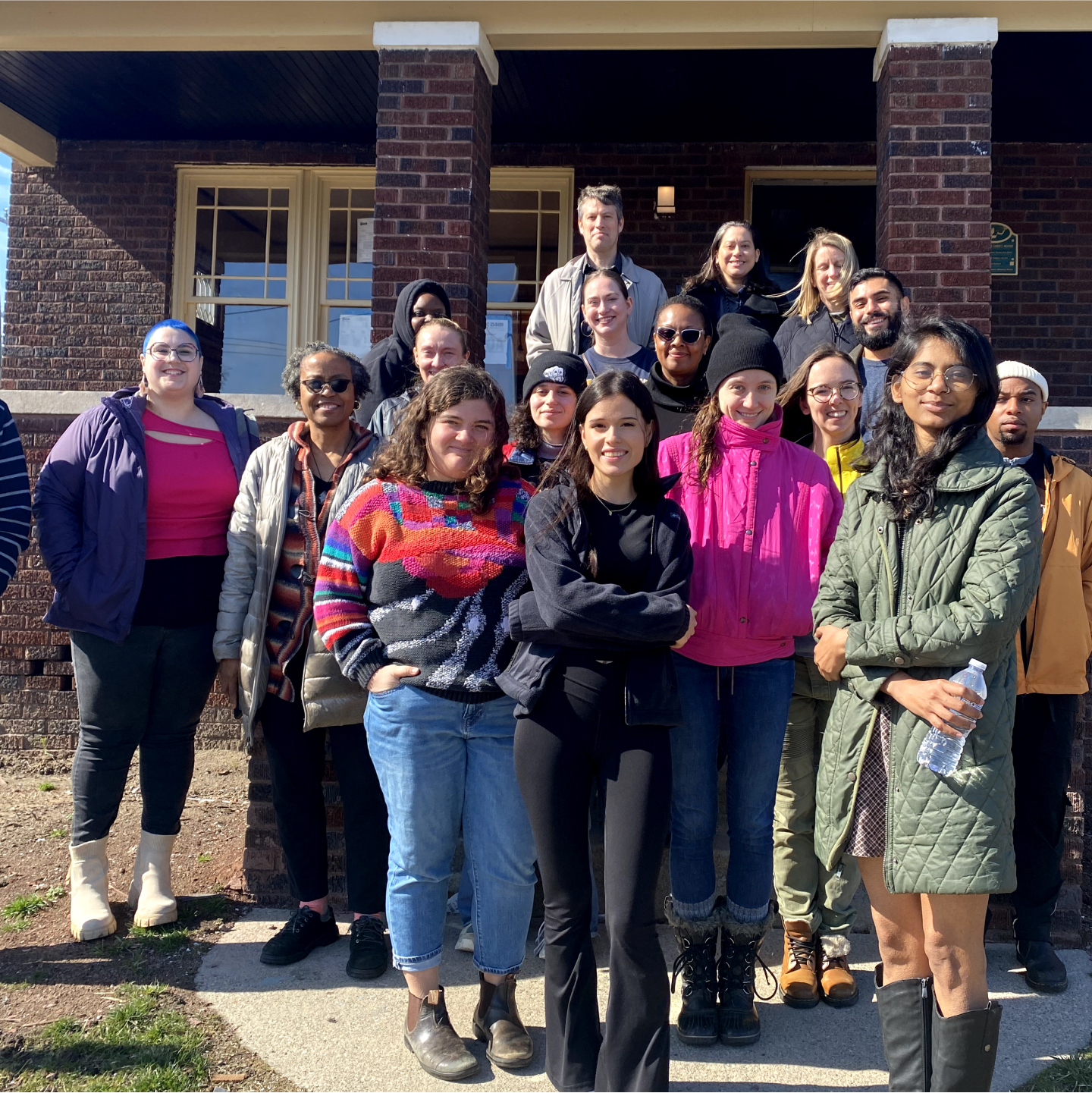 A group of people poses for a photo outside a brick building on a sunny day. Some are standing on the sidewalk and others are on the porch steps.