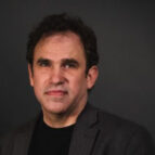 A man with short curly hair wearing a dark blazer and black shirt poses against a plain dark background.