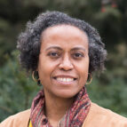 A person with short curly hair, gold hoop earrings, and a patterned scarf smiles outdoors with trees in the background.