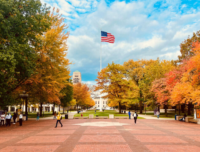 People walk across a brick plaza lined with autumn trees. An American flag flies on a tall pole, with a domed building and clock tower in the background under a partly cloudy sky.