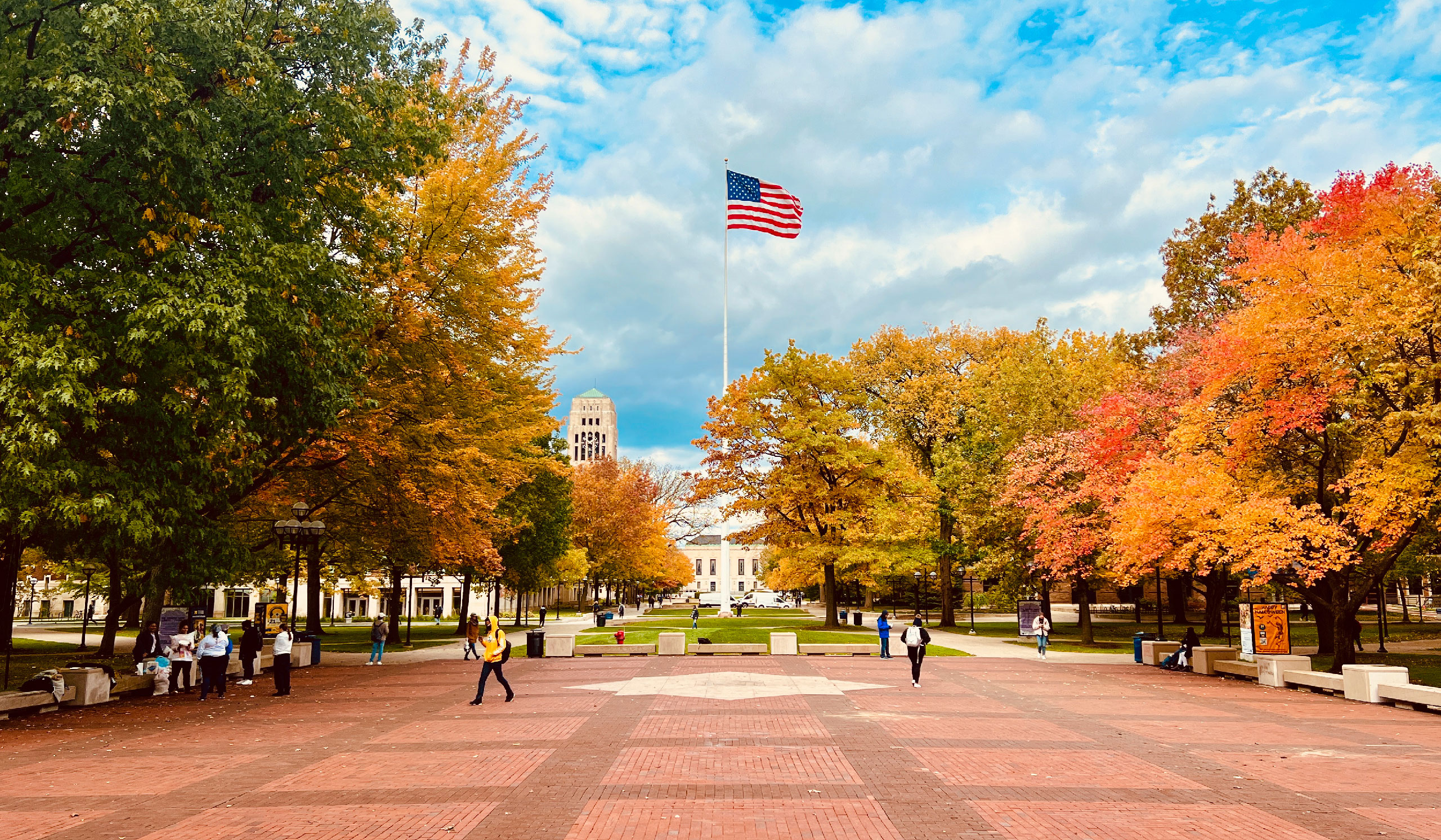 People walk across a brick plaza lined with autumn trees. An American flag flies on a tall pole, with a domed building and clock tower in the background under a partly cloudy sky.