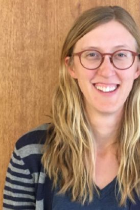 A woman with long blonde hair and glasses, wearing a striped sweater over a blue shirt, stands in front of a wooden background, smiling at the camera.