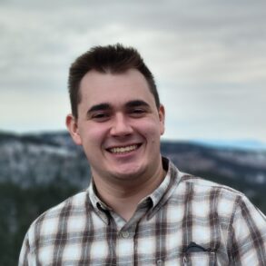 A man in a plaid shirt smiles outdoors with a blurred, snowy landscape in the background.