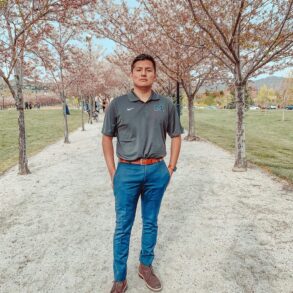 A man stands on a gravel path lined with blooming trees in a park, wearing a dark polo shirt, blue pants, and brown shoes, with his hands in his pockets.