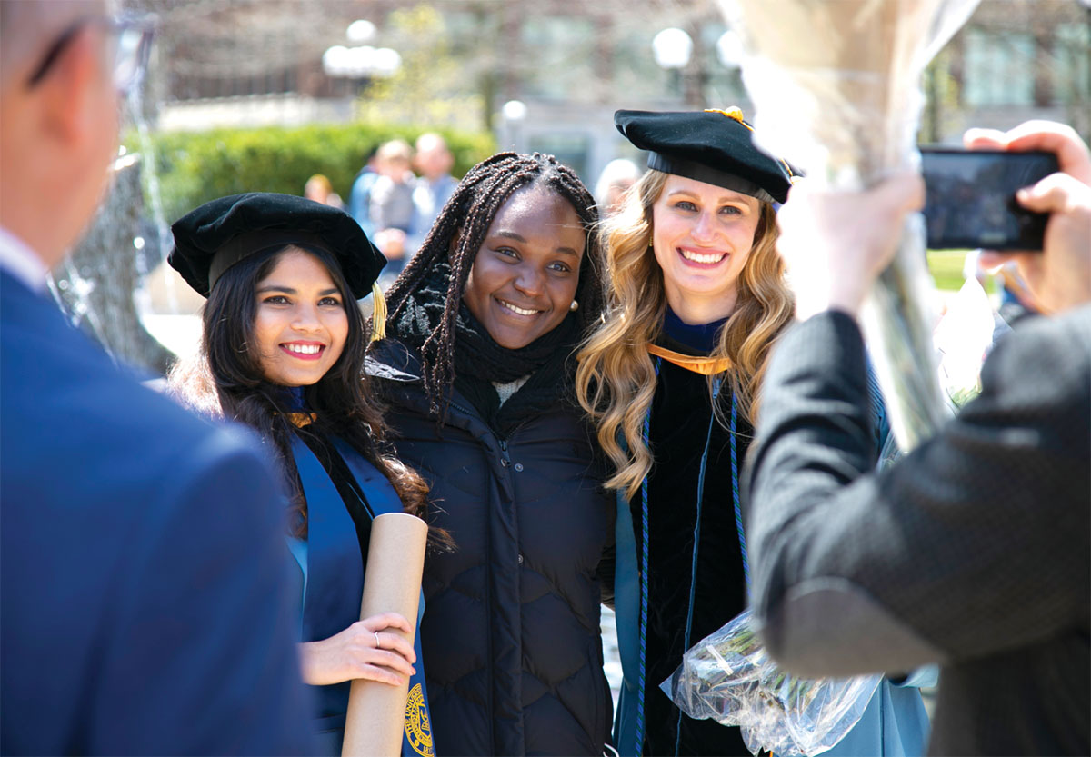 Three women pose and smile for a photo at a graduation ceremony, with one holding a diploma and another holding flowers.