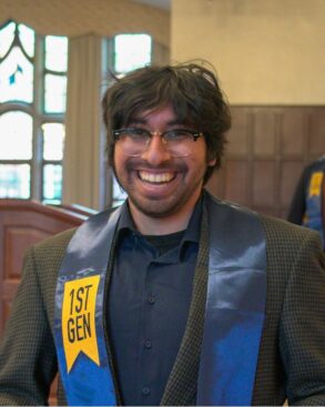 A person wearing glasses and a "1st GEN" sash smiles indoors, standing in front of wood paneling and large windows.