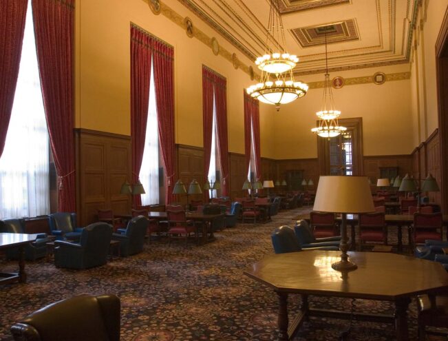 A spacious room with high ceilings, large red drapes, chandeliers, wood-paneled walls, and various tables and armchairs arranged throughout.