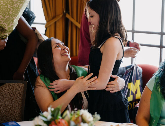 A smiling woman sitting at a table embraces a young girl standing beside her; other people and a bouquet of flowers are visible in the background.