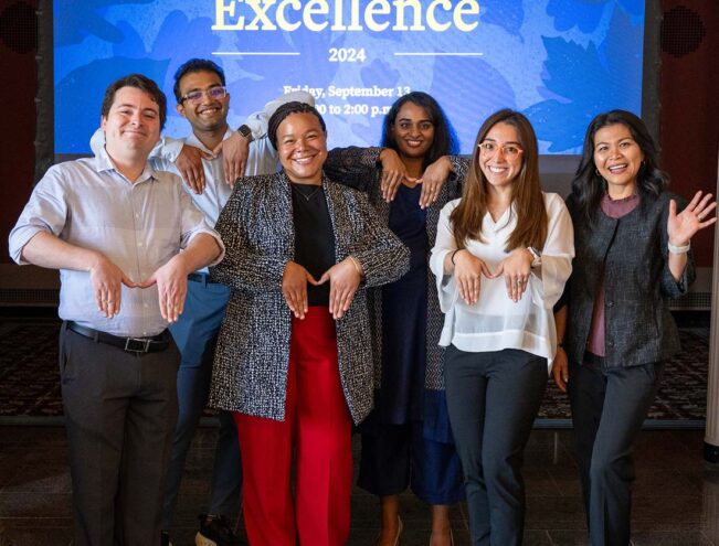 Six people stand in front of an "Excellence 2024" event sign, smiling and posing with their hands bent at the wrists, imitating animal paws.