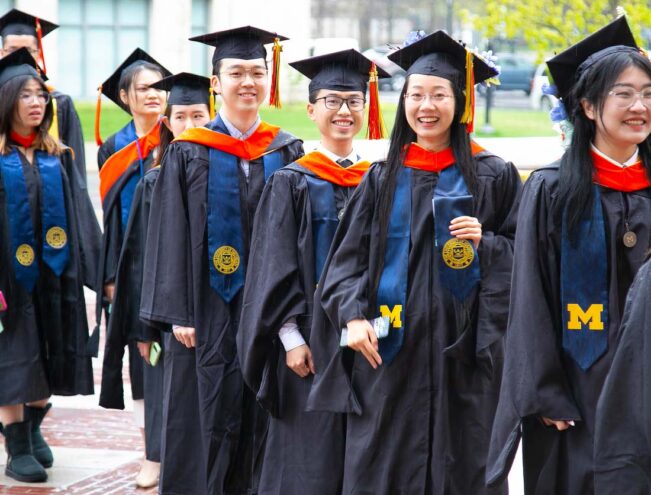 A group of graduates in caps and gowns, wearing blue and orange academic stoles, walk together at a university graduation ceremony.