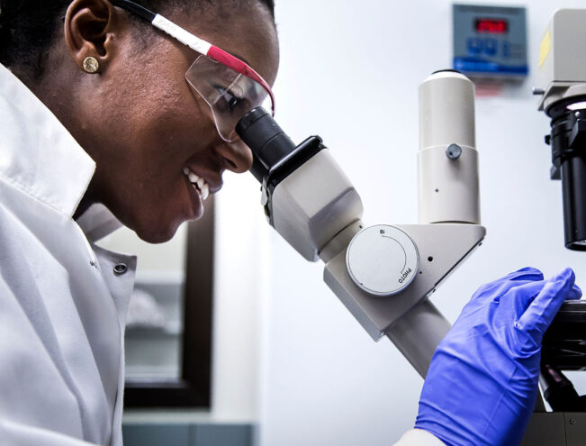 A person wearing safety glasses and gloves looks into a microscope in a laboratory setting.