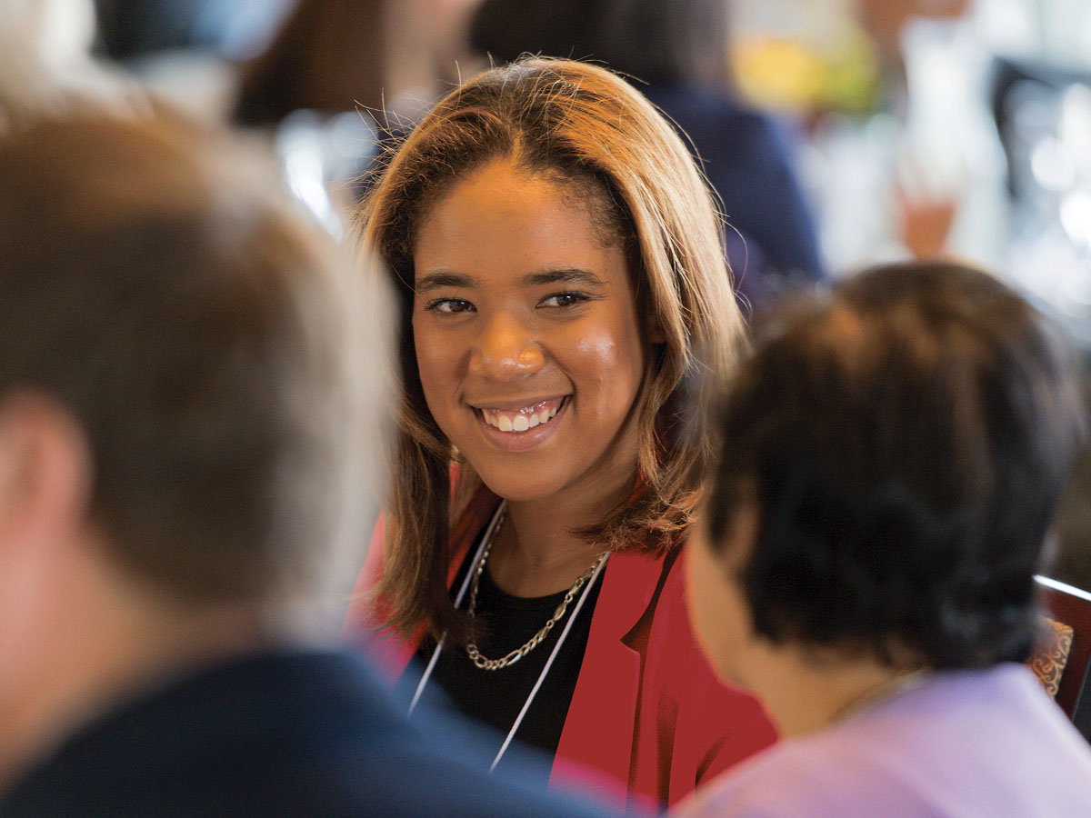 A woman with light brown hair smiles while seated at a table with other people in a social or professional setting.