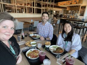 Three people sit at a restaurant table with various dishes, drinks, and chips. They are smiling at the camera in a modern, well-lit dining area.