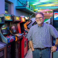 A man stands and smiles in front of a row of vintage arcade game machines in a brightly lit room.