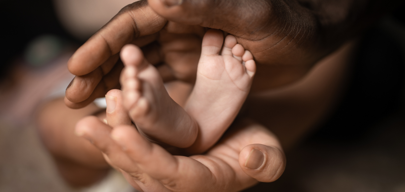 Close-up of two adult hands gently holding a baby's feet, highlighting contrast in skin tones and focus on tenderness and protection.