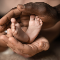 Close-up of two adult hands gently holding a baby's feet, highlighting contrast in skin tones and focus on tenderness and protection.