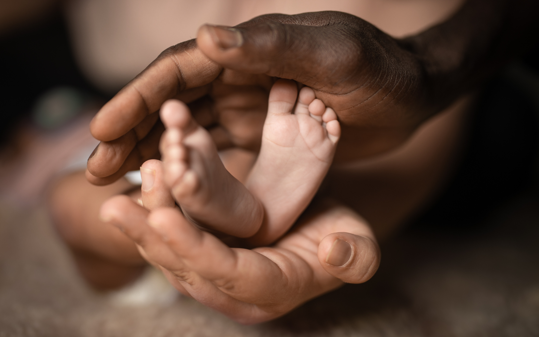 Close-up of two adult hands gently holding a baby's feet, highlighting contrast in skin tones and focus on tenderness and protection.