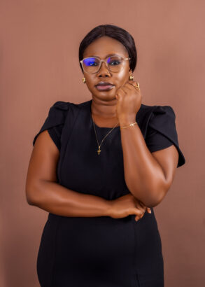 A woman wearing glasses and a black dress stands against a brown background, resting her left arm on her right and touching her face with her right hand.