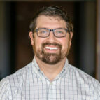 Man with glasses, beard, and mustache wearing a white checkered shirt, smiling at the camera against a blurred indoor background.