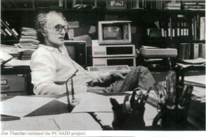 An older man with curly hair and glasses sits at a cluttered desk in an office with papers, books, and a vintage computer behind him.